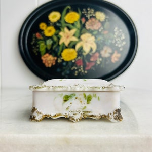 Op de afbeelding: Een klein wit porseleinen sieradendoosje met een bloemenmotief en gouden accenten. Het doosje heeft een scharnierend deksel en staat op vier sierlijke pootjes.