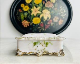 May include: A small white porcelain trinket box with a floral design and gold accents. The box has a hinged lid and sits on four ornate feet.