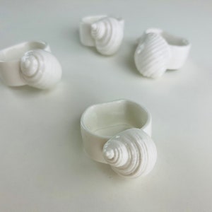 May include: Four white ceramic rings shaped like seashells. The rings are all different sizes and shapes, and they are all made of a smooth, white ceramic.