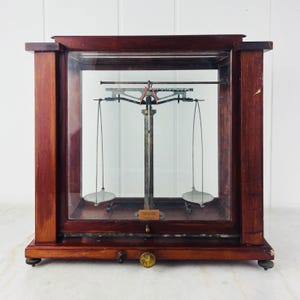 May include: An antique analytical balance scale enclosed in a dark wood and glass case. The internal mechanism features a central metal pillar with a horizontal beam and two suspended circular weighing pans. A small brass knob is on the front base.