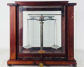 May include: An antique analytical balance scale enclosed in a dark wood and glass case. The internal mechanism features a central metal pillar with a horizontal beam and two suspended circular weighing pans. A small brass knob is on the front base.