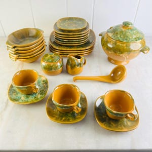 May include: A vintage ceramic dinner service in yellow and green hues. The set comprises bowls, plates, cups, saucers, a sugar bowl, a creamer, a lidded soup tureen, and a serving spoon. The items feature a speckled glaze finish.