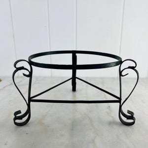 May include: Black metal plant stand with a circular top and decorative heart-shaped accents on the sides. The stand has a three-legged base and is designed to hold a potted plant or decorative item.