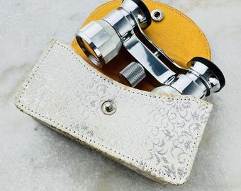 May include: Silver opera glasses with a white and silver patterned case. The case has a yellow lining.