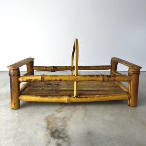 May include: A rectangular bamboo basket featuring a slatted base and a central arched handle. The structure is constructed from natural light brown bamboo stalks with darker brown nodes and joints, forming a sturdy frame with four corner posts.