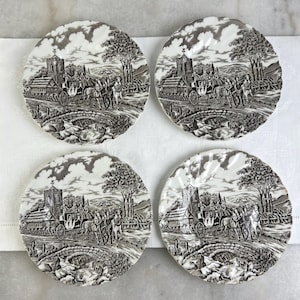 Può includere: Quattro piatti decorativi coordinati con un design transferware in bianco e nero. Ogni piatto presenta un paesaggio rurale con una carrozza trainata da cavalli, un ponte ed edifici. I piatti sono disposti su una superficie bianca.