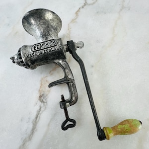 May include: Vintage metal meat grinder with a hand crank. The grinder is silver with black accents and a wooden handle with green paint. The text "BEATRICE MADE IN ENGLAND" is visible on the side of the grinder.