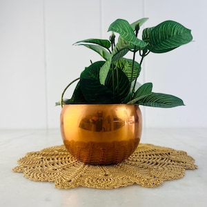 May include: A shiny copper-colored planter with a small handle, holding a green plant with large, veined leaves. The planter sits on a gold-colored, crocheted doily with a scalloped edge. The background is a white wall.