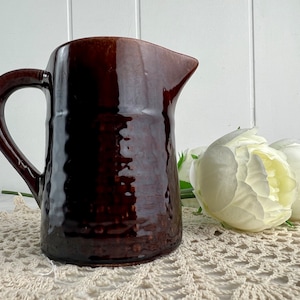 May include: A dark brown ceramic jug with a glossy, dimpled texture, a curved handle, and a prominent pouring spout. The jug features a cylindrical body with a slightly uneven, hand-crafted appearance and a deep, reflective glaze finish.