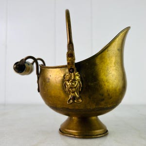 May include: A vintage brass coal scuttle with a handle and a decorative lion's head. The scuttle has a curved spout and a small, round base. The handle has a ceramic grip with blue and white floral details.