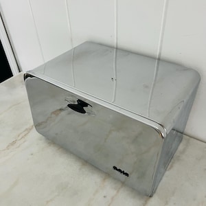 Op de afbeelding: Een glanzende, verchroomde metalen doos met een gebogen bovenkant en een kleine, zwarte handgreep. De doos heeft een rechthoekige vorm en staat op een marmeren oppervlak. Het woord "Raim" staat op de voorkant.