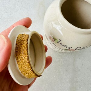 K&ouml;nnte beinhalten: Wei&szlig;e Keramikdose mit Korkdeckel und Blumenmuster. Die Dose tr&auml;gt die Aufschrift "Instant Coffee" auf der Vorderseite.