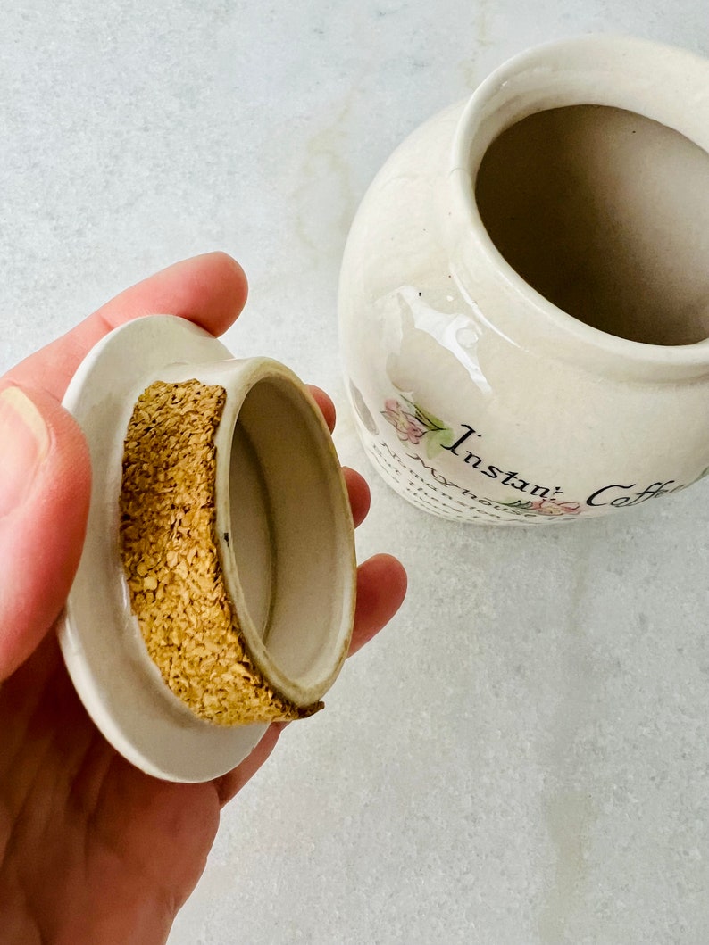 K&ouml;nnte beinhalten: Wei&szlig;e Keramikdose mit Korkdeckel und Blumenmuster. Die Dose tr&auml;gt die Aufschrift "Instant Coffee" auf der Vorderseite.