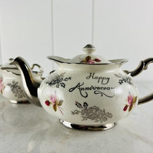 Peut inclure: Un ensemble théière et crémier blanc avec des accents argentés et des motifs floraux. La théière a une poignée et un bec verseur, et le crémier a un couvercle. La théière porte l'inscription "Happy Anniversary".