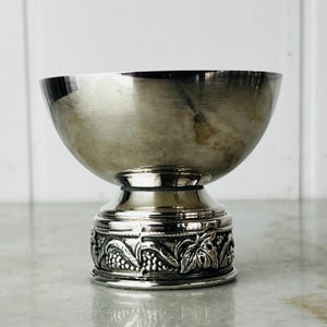 May include: A polished silver-toned metal bowl with a smooth, reflective interior and a wide, flared rim. The base features a decorative band with a raised relief pattern of grape clusters and vine leaves, providing a textured contrast to the bowl.