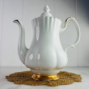 May include: A white porcelain coffee pot with gold detailing on the base and handle. The pot features a fluted design and a decorative lid. It rests on a gold crocheted doily. The spout and handle have elegant curves. A classic design.