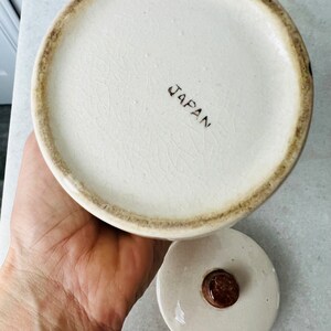 K&ouml;nnte beinhalten: Wei&szlig;e Keramikdose mit braunem Deckel und braunem Knopf. Auf dem Boden der Dose steht "JAPAN" gedruckt.