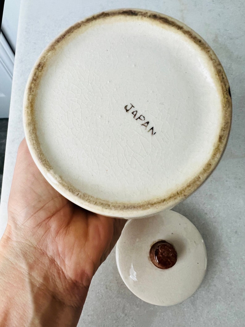 K&ouml;nnte beinhalten: Wei&szlig;e Keramikdose mit braunem Deckel und braunem Knopf. Auf dem Boden der Dose steht "JAPAN" gedruckt.