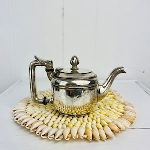 May include: A silver teapot with an ornate handle and lid sits on a round coaster made of white seashells.