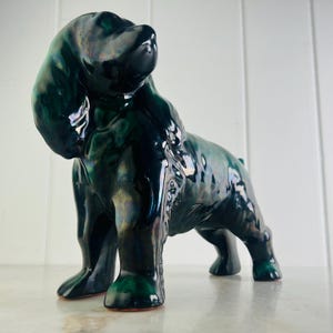 以下が含まれることがあります： 光沢のあるイリデセントダークグリーンの釉薬を施したセラミック製の犬の置物。犬は立っており、頭を上に向けています。この置物は詳細な特徴と滑らかで反射する表面を持っています。装飾品。