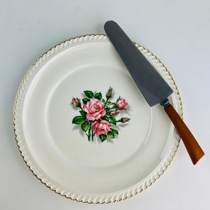 Pode incluir: Um prato de bolo branco com uma borda dourada e um desenho floral com rosas cor-de-rosa. Uma faca de bolo prateada com uma pega castanha é colocada no prato.
