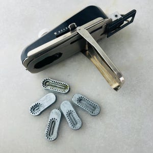 Könnte beinhalten: Ein schwarzes und silbernes Vintage-Etikettiergerät mit dem Text "W 65432" und mehreren silbernen Etikettenkartuschen. Das Etikettiergerät hat einen silbernen Griff und einen weißen Knopf. Die Kartuschen sind oval geformt und haben ein geriffeltes Design.