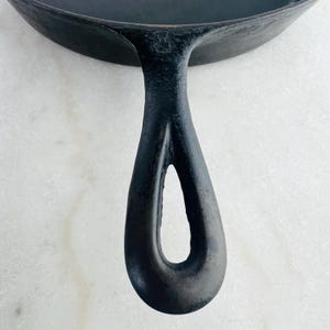May include: Close-up of a black cast iron skillet with a long, curved handle. The handle has a teardrop-shaped opening for hanging. The skillet's surface is dark and textured, suggesting it's well-seasoned and ready for cooking.