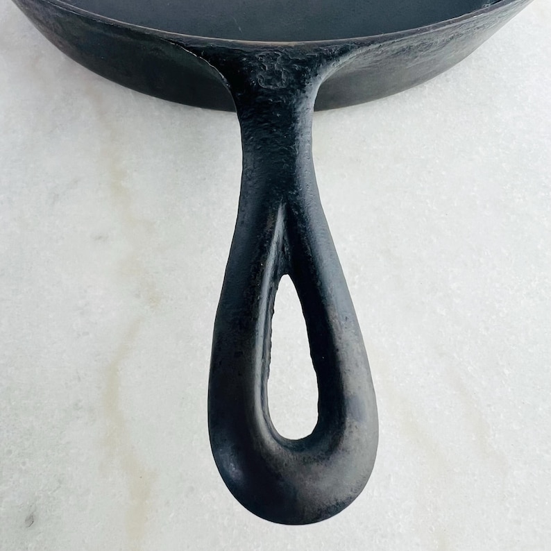 May include: Close-up of a black cast iron skillet with a long, curved handle. The handle has a teardrop-shaped opening for hanging. The skillet's surface is dark and textured, suggesting it's well-seasoned and ready for cooking.