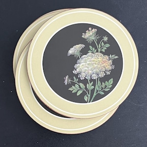 May include: Set of three round coasters. Each coaster features a black center with a floral design in white, yellow, and green. The coasters have a light beige border with a thin white and brown accent ring.