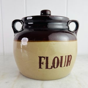 Puede incluir: Un tarro de harina de cerámica vintage con tapa y asas de color marrón oscuro. El tarro tiene una base beige con la palabra "FLOUR" en letras marrones. El tarro está diseñado para el almacenamiento en la cocina.