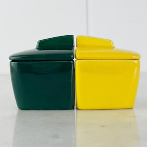 May include: Two ceramic lidded containers, one green and one yellow. The containers are square with rounded edges and have lids that fit snugly on top.