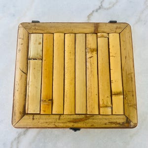 May include: A square bamboo box with a hinged lid. The lid is made of vertical bamboo strips, and the frame is a darker shade of bamboo. The box is closed with a small metal clasp.