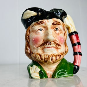 Op de afbeelding: Een keramische mok in de vorm van een mannenhoofd met een zwarte hoed, een groen jasje en een rood-zwarte gestreepte handgreep. De man heeft een bruine baard en snor, en zijn gezicht is geschilderd met realistische details.