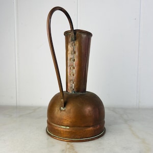 Puede incluir: Una jarra de cobre vintage con base redondeada, cuello alto y cilíndrico y asa curva. La jarra tiene un tono marrón rojizo cálido y muestra signos de edad y uso. El asa está unida al cuello y a la base.