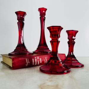 May include: Four ruby red glass candle holders of varying heights arranged on a red book titled Something's Got to Give by Hargrove Sloane. The candle holders feature a faceted, multi-sided base and stem design with a wide, flared top rim.