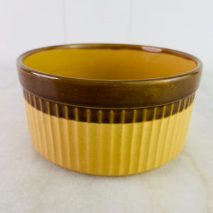 Puede incluir: Un ramequín redondo de cerámica con exterior acanalado e interior liso. El borde es de color marrón oscuro, contrastando con el interior y el exterior amarillo dorado. El ramequín está diseñado para hornear y servir porciones individuales.