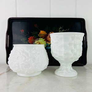 May include: Two white milk glass vases with a raised floral design. The vases are sitting on a black tray with a floral design.