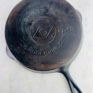 May include: A well-worn, cast iron skillet with a long handle and a circular cooking surface. The bottom of the pan features the embossed text "SMART" above a diamond-shaped logo and the text "BROCKVILLE K-4".