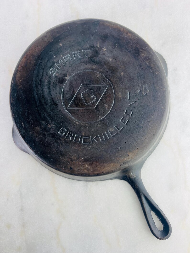 May include: A well-worn, cast iron skillet with a long handle and a circular cooking surface. The bottom of the pan features the embossed text "SMART" above a diamond-shaped logo and the text "BROCKVILLE K-4".