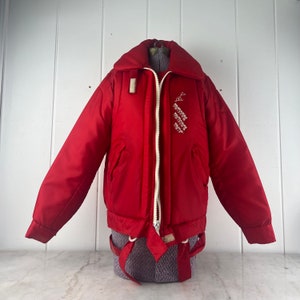 May include: Red puffer jacket with a white zipper and a white embroidered logo on the right side. The jacket has a drawstring waist and a white zipper pull.