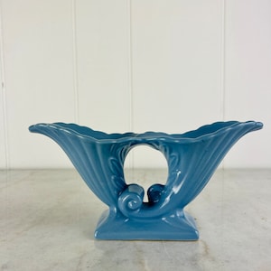 Könnte beinhalten: Eine hellblaue Keramikvase mit einem einzigartigen, geschwungenen Design. Die Vase hat eine breite, offene Oberseite und einen schmalen Boden.