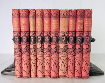 Könnte beinhalten: Eine Sammlung antiker Bücher mit korallenfarbenen Einbänden. Die Buchrücken zeigen goldene Schrift und ein Porträt eines Mannes. Die Bücher sind in einer Reihe angeordnet und werden von einem dekorativen Buchstützen gehalten.
