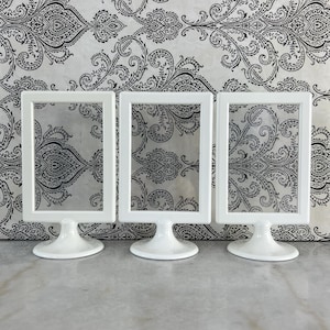 May include: Three white rectangular picture frames with a pedestal base. The frames have a clear center, and are set against a black and white patterned background. The frames are suitable for displaying photos or artwork.