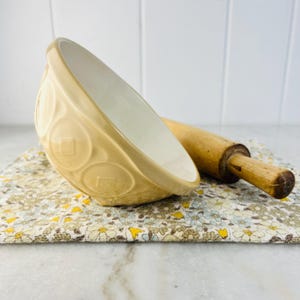 Puede incluir: Un cuenco de cerámica beige con interior blanco y diseños circulares en relieve. Un rodillo de madera descansa junto al cuenco sobre un paño con estampado floral. Ideal para hornear y cocinar.