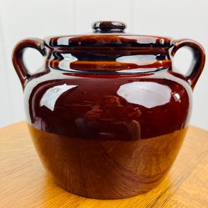 Könnte beinhalten: Ein brauner Keramik-Bohnentopf mit Deckel und zwei Griffen. Der Topf hat eine glänzende Oberfläche.