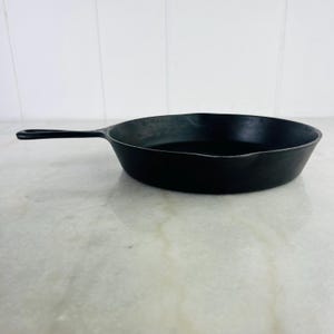 May include: A black cast iron skillet with a long handle. The skillet is round with a slightly curved rim and is sitting on a white and gray surface. The handle has a hole at the end for hanging.