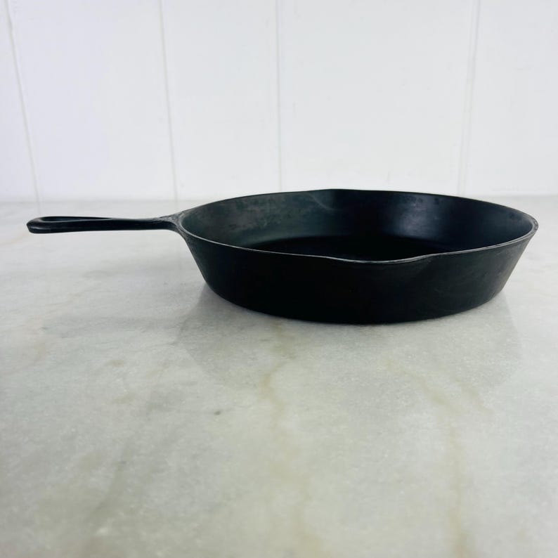 May include: A black cast iron skillet with a long handle. The skillet is round with a slightly curved rim and is sitting on a white and gray surface. The handle has a hole at the end for hanging.