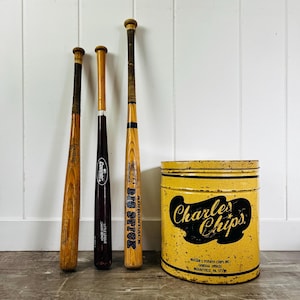 Może przedstawiać: Trzy drewniane kije baseballowe w różnych odcieniach i markach, w tym Louisville i Adirondack, stojące pionowo obok cylindrycznej żółtej metalowej puszki Charles Chips z czarnym napisem i detalami z odpryskującą farbą.