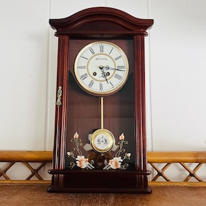 May include: A dark wood wall clock with a cream-colored face and black Roman numerals. The clock has a pendulum and floral accents. The clock face reads "Westminster". The clock is rectangular with a curved top.