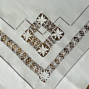 Peut inclure: Gros plan sur une nappe blanche avec des détails de dentelle complexes. Le motif présente des formes géométriques et des motifs floraux, avec un mélange de tissu uni et de dentelle ajourée. Les bords sont finis avec une bordure délicate cousue.
